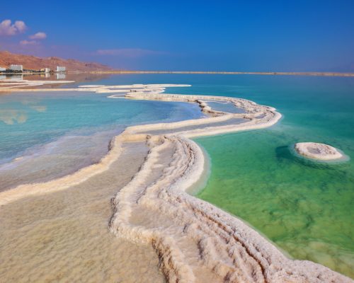 The path from salt picturesquely curls in salty water. Hotels are reflected in smooth water ashore. Israeli coast of the Dead Sea